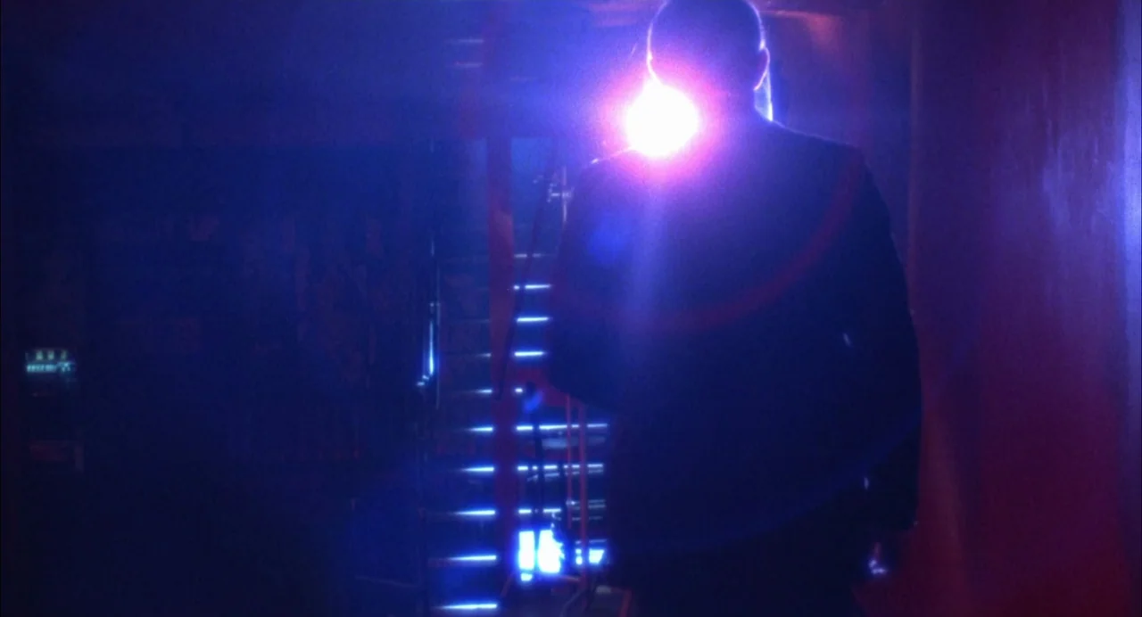 The Killing of a Chinese Bookie (1976) movie still: medium — A dimly lit industrial stairwell glows with intense blue and red light sources. A man…