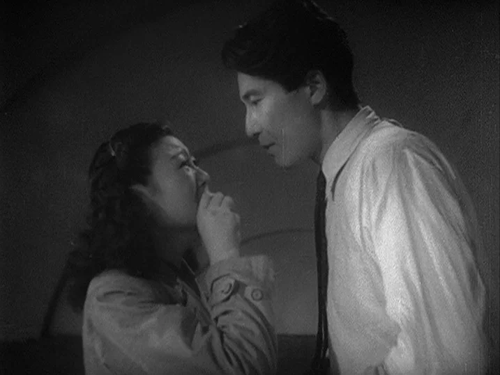 One Wonderful Sunday (1947) movie still: medium — An dimly lit interior space with shadows cast across the wall. A man stands over a woman who holds…