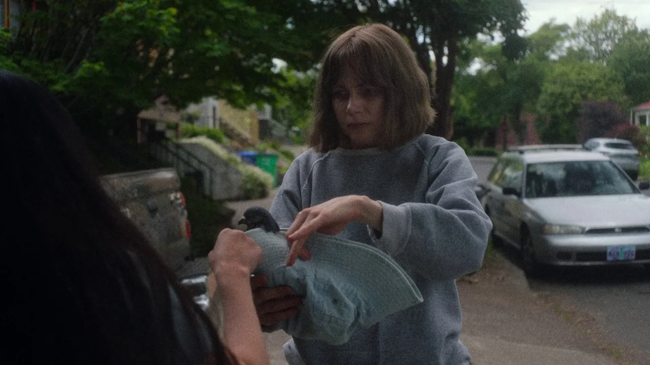 Showing Up (2022) movie still: medium — A quiet suburban street with trees and a parked silver car in the background. A woman holds a small bird…