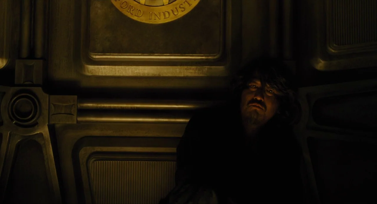 Snowpiercer (2013) movie still: medium — A man sits against a metallic wall featuring an embossed circular industry logo. The room is dimly lit and…