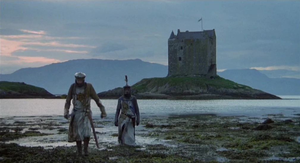 Monty Python and the Holy Grail (1975) movie still: fullbody — A rocky tidal shoreline features a stone tower on a small island background. Two…