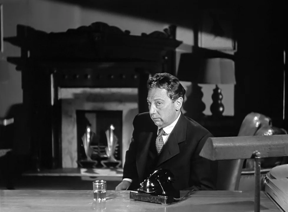 M (1951) (1951) movie still: medium — A dark office features a large fireplace and a desk with a vintage telephone. A man in a dark suit sits at the…