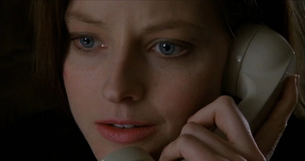 The Silence Of The Lambs (1991) movie still: closeup — A close view of a woman holding a vintage telephone receiver to her ear. Dark, out-of-focus…