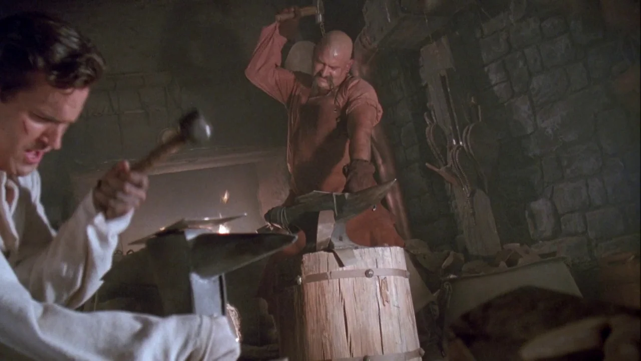 Army of Darkness (1992) movie still: medium — A dimly lit stone blacksmith shop features iron tools and a burning forge. A muscular man strikes…