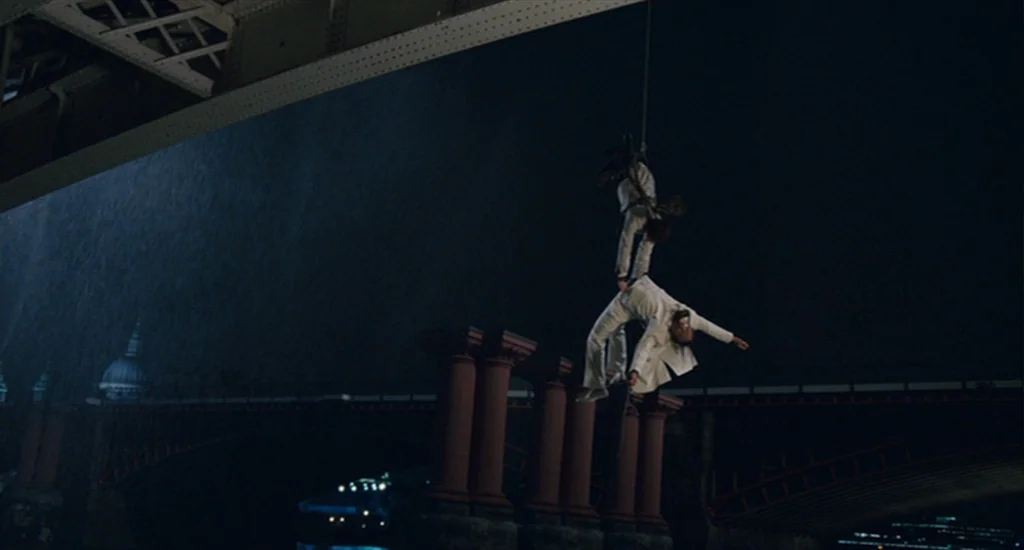 The Imaginarium of Doctor Parnassus (2009) movie still: wide — Two individuals hang from a rope beneath the span of a large iron bridge. The…