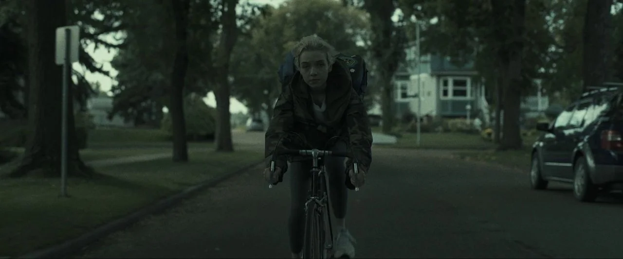 Come True (2020) movie still: medium — A suburban street lined with mature trees and detached houses during an overcast day. A young girl rides a…
