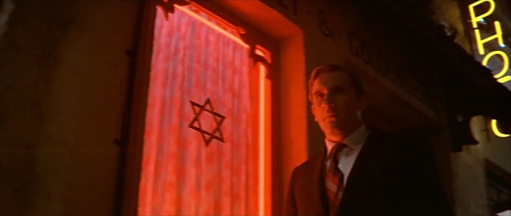 The Moon in the Gutter (1983) movie still: medium — An exterior city street at night is illuminated by saturated red neon light. A man in a dark…