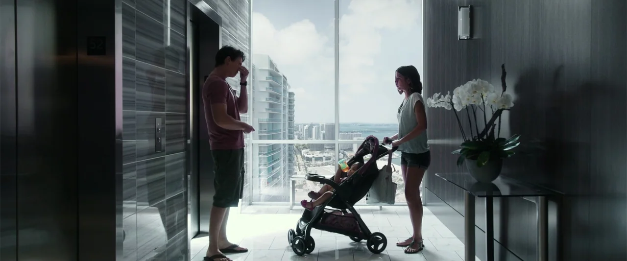 War Dogs (2016) movie still: wide — A high-rise apartment hallway features wood-paneled walls, an elevator bank, and floor-to-ceiling windows. A man…
