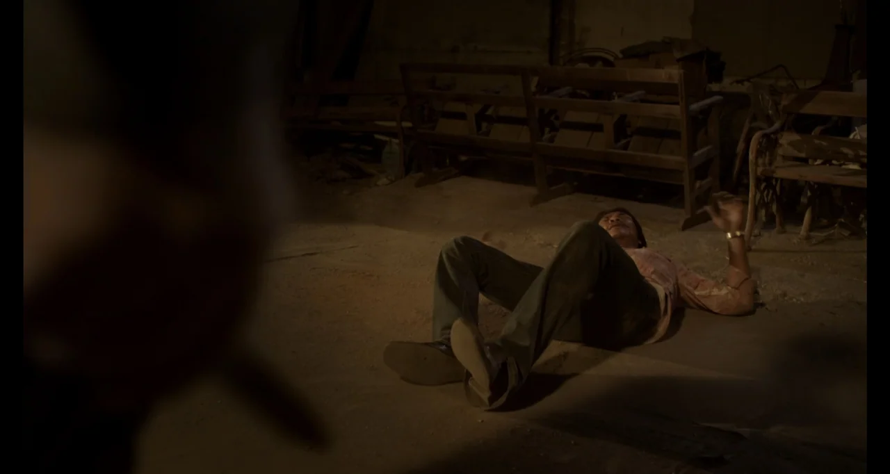 Ninja: Shadow of a Tear (2013) movie still: wide — A dark, dusty barn interior features wooden benches in the background. A man lies prone on the…