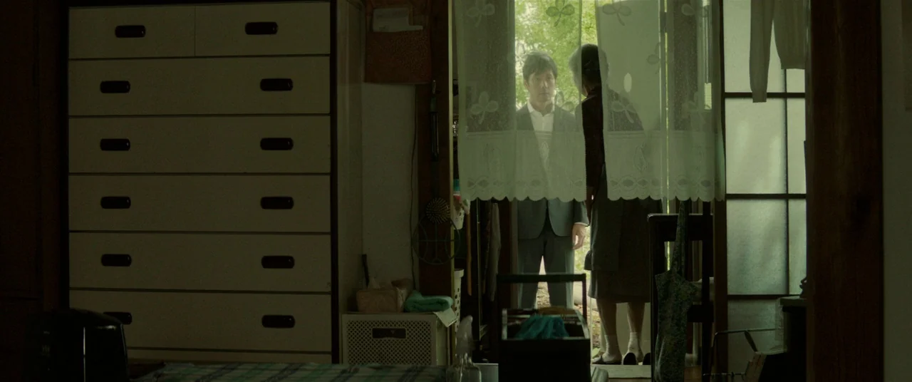 Creepy (2016) movie still: medium — A dimly lit room with a large chest of drawers sits in the foreground. A man and a woman stand by an open door…