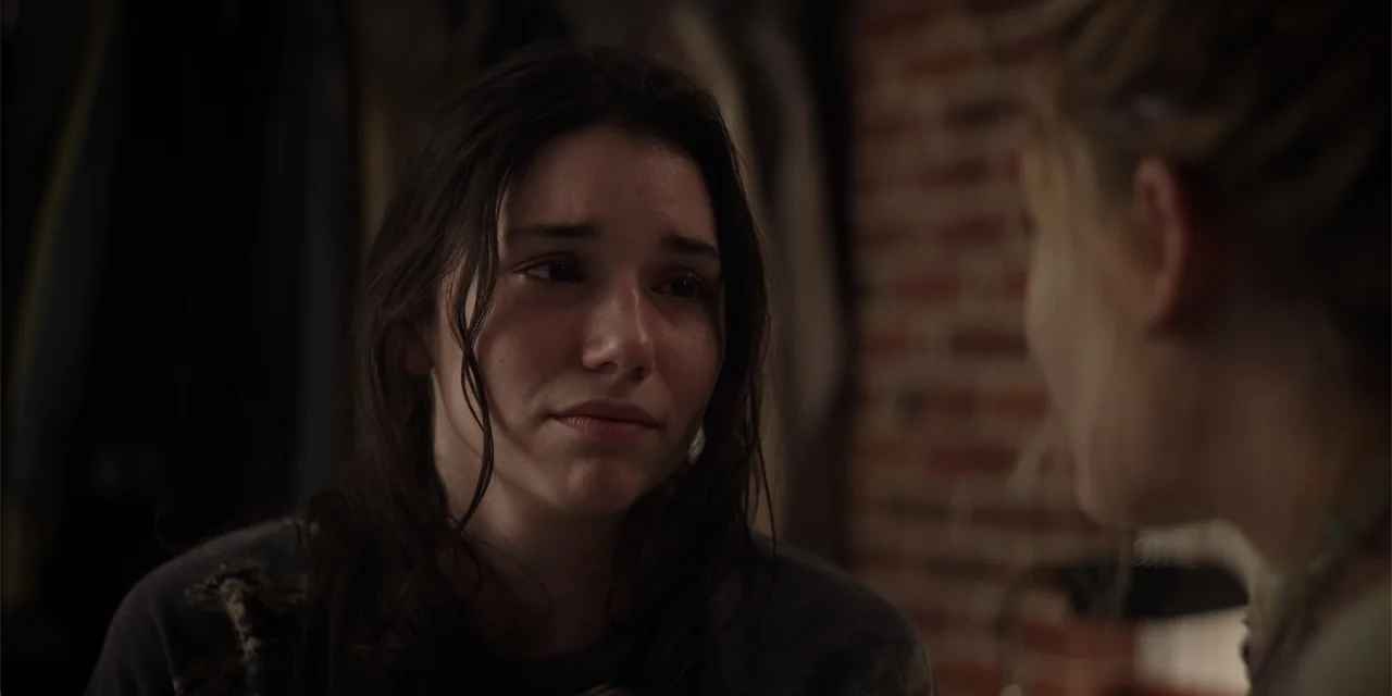 Fall (2022) movie still: closeup — A dimly lit interior room features a blurred brick wall in the background. A young woman with dark hair looks…