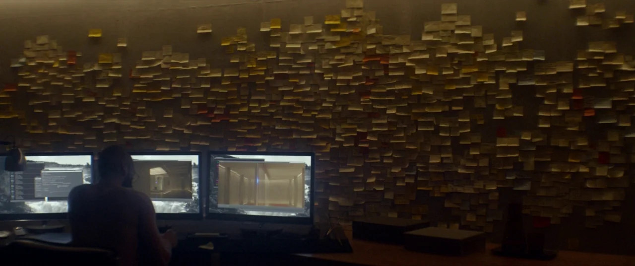 Ex Machina (2015) movie still: wide — A dimly lit room features a massive wall densely covered in small sticky notes. A man sits at a desk in front…