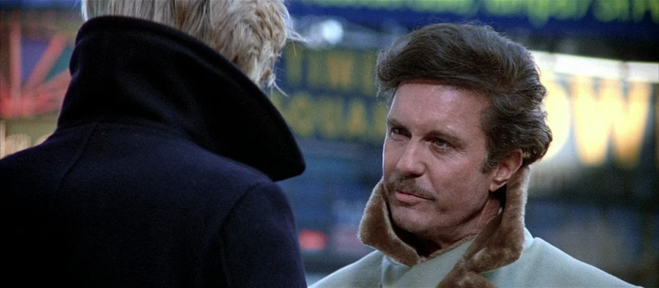 Three Days of the Condor (1975) movie still: medium — A night street scene with blurred neon lights in the background. A man faces an unseen person…