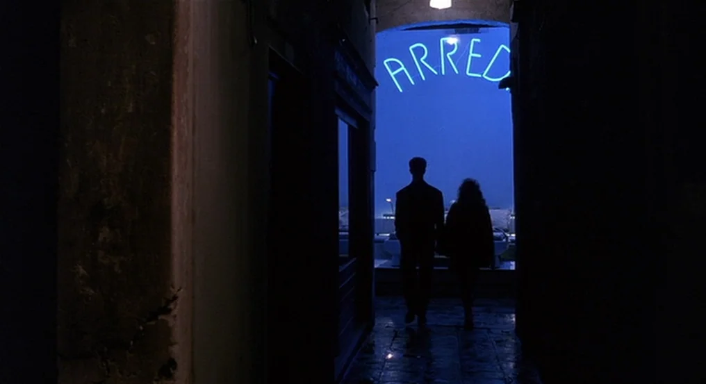 The Comfort of Strangers (1990) movie still: fullbody — A narrow stone passageway frames a distant view of a night harbor. A man and a woman walk…