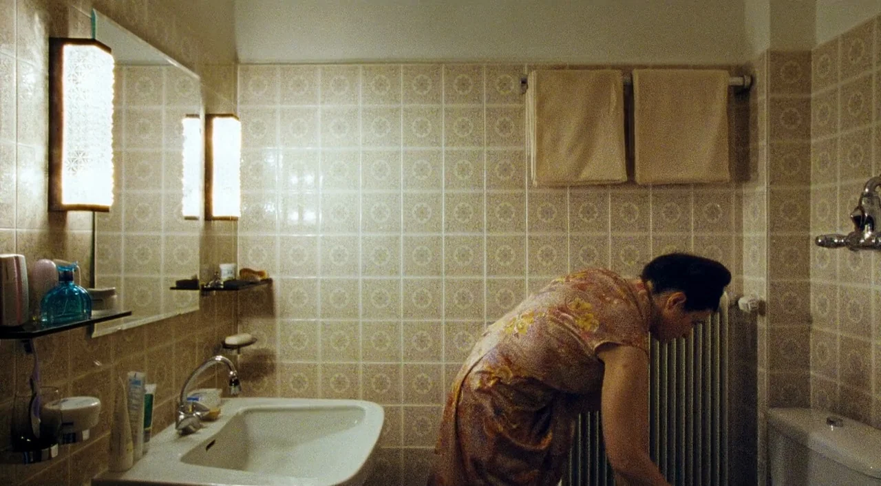 Paradise: Faith (2012) movie still: medium — A cramped tiled bathroom features floral patterned wall tiles and two beige hanging towels. A woman…