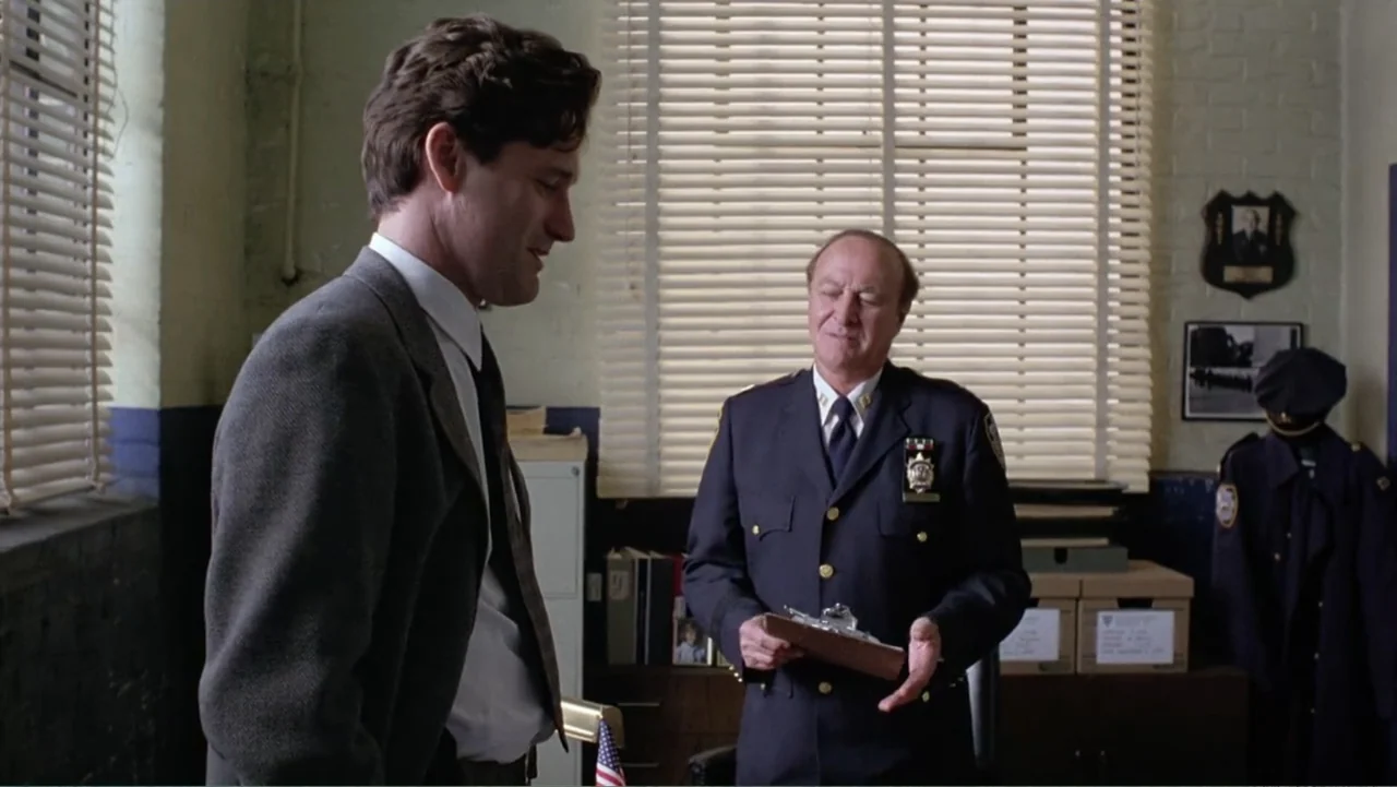 Mistrial (1996) movie still: medium — A municipal office features a desk, stacks of files, and a wall-mounted police shield. A man in a suit speaks…