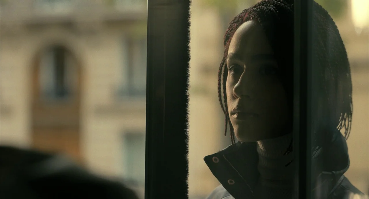 The Killer (2024) (2024) movie still: closeup — A woman stands behind a glass pane at a city street bus stop. She looks through the window while the…