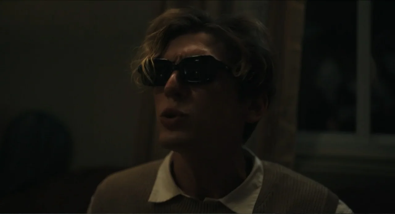 Hell of a Summer (2025) movie still: closeup — A dimly lit room interior with a window visible in the background shadow. A young man wearing dark…