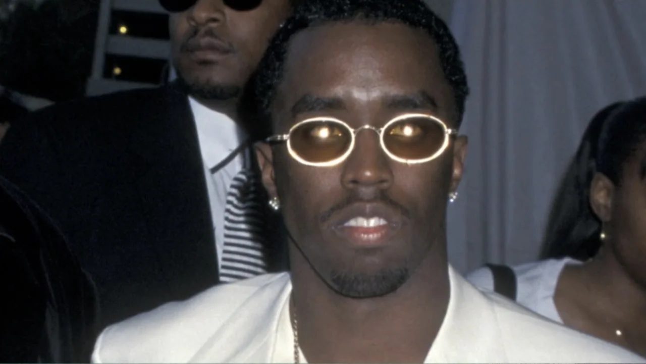 Diddy: The Making of a Bad Boy (2025) movie still: closeup — A public event setting with out-of-focus figures in the background near a white…