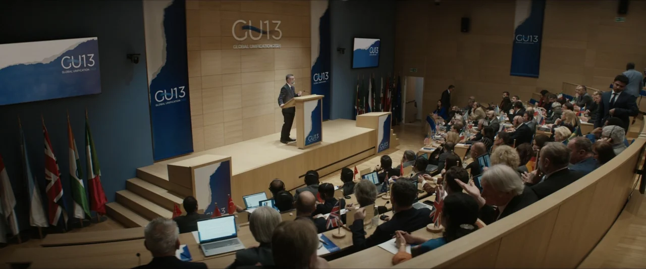 Canary Black (2024) movie still: wide — A modern conference hall features wooden tiered seating facing a central podium stage. A man stands at the…