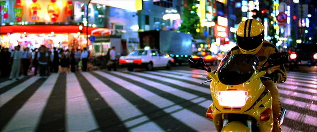Kill Bill Vol. 1 (2004) movie still: medium — An urban pedestrian crossing at night is illuminated by vibrant neon signage and streetlights. A…