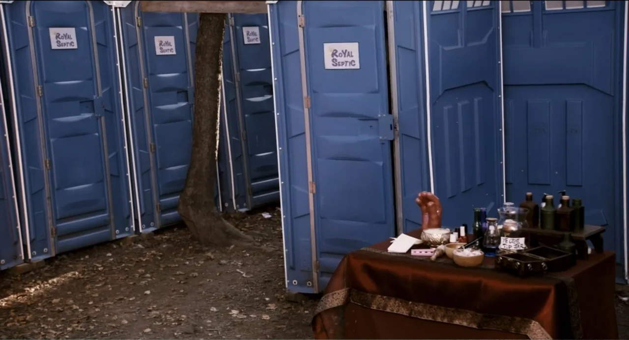 Todo es amor (2024) movie still: wide — A row of blue portable toilets stands on a patch of dirt and fallen leaves. In the foreground a table with a…