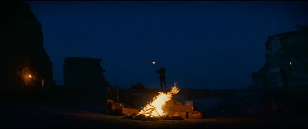 The Darjeeling Limited (2007) movie still: wide — A dark, expansive quarry at night illuminated only by a large central bonfire. Three silhouetted…