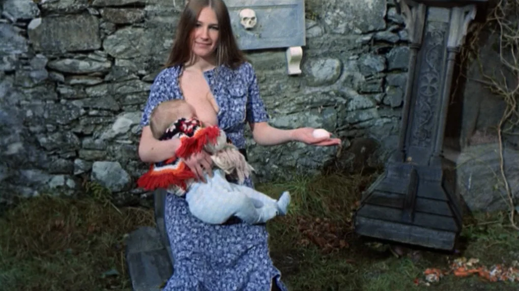The Wicker Man movie still: medium — A woman stands before a weathered stone wall with a small skull ornament. She holds an infant in her arms and…