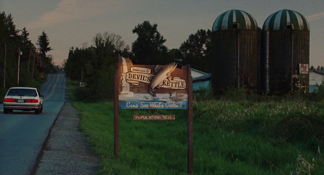 Jennifer’s Body (2009) movie still: wide — A rural asphalt road runs past a wooden town sign and two silos. A white car travels away from the camera…