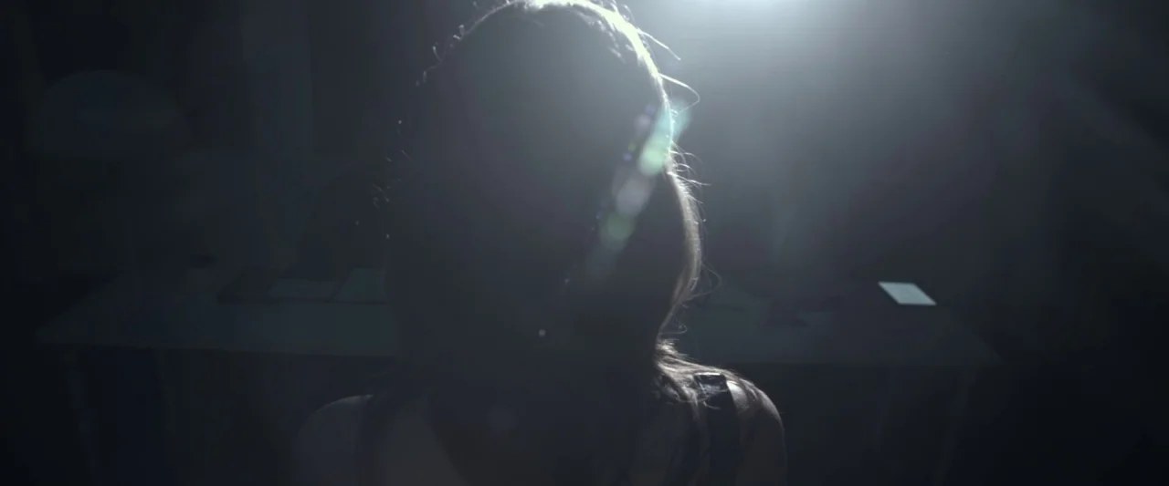 Starry Eyes (2014) movie still: medium — A person sits in a dark space in front of a white table surface. The figure is obscured by heavy backlight…