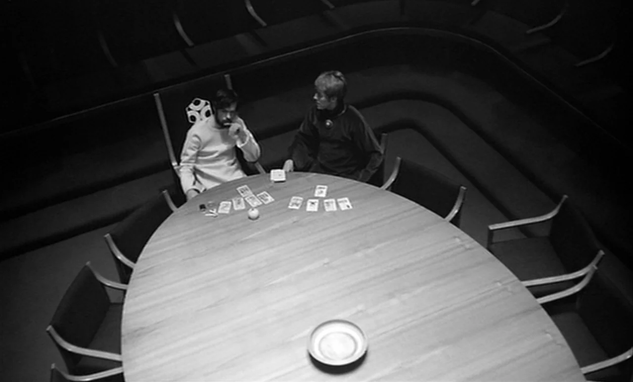 Stereo (1969) movie still: wide — A modernist room features a large oval wooden table surrounded by curved built-in seating. Two men sit at the…