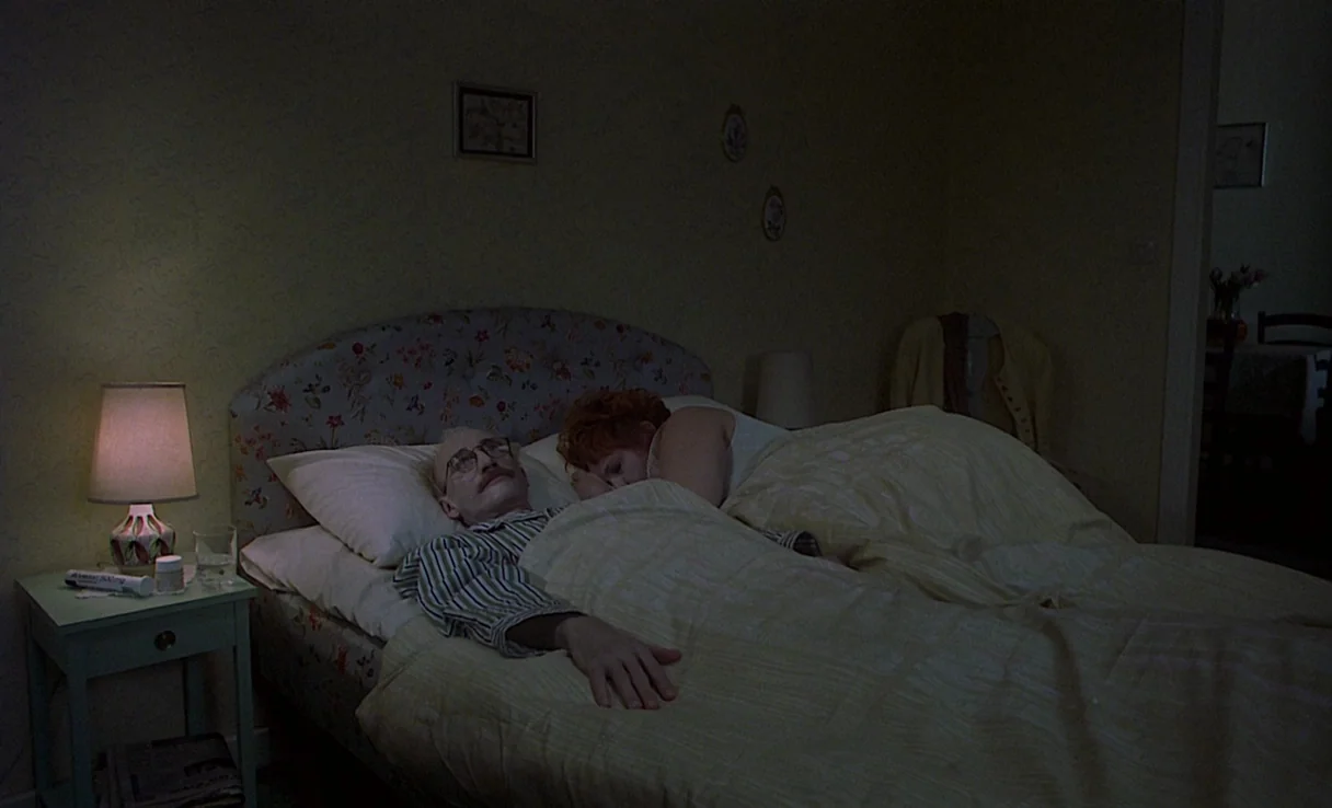 Songs from the Second Floor (2000) movie still: medium — An older couple lies in bed within a dimly lit room featuring floral wallpaper. The man…