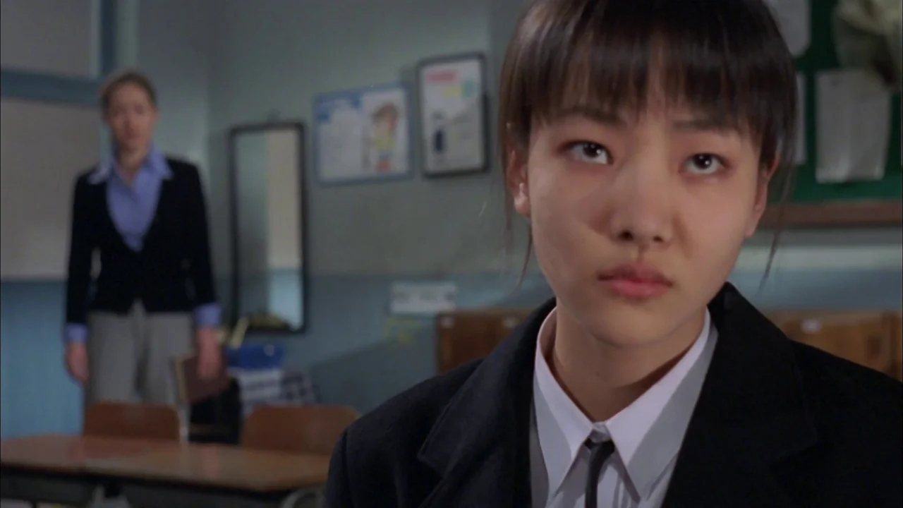 Whispering Corridors (1998) movie still: closeup — A school classroom featuring desks, chairs, and framed papers hanging on the back wall. A student…