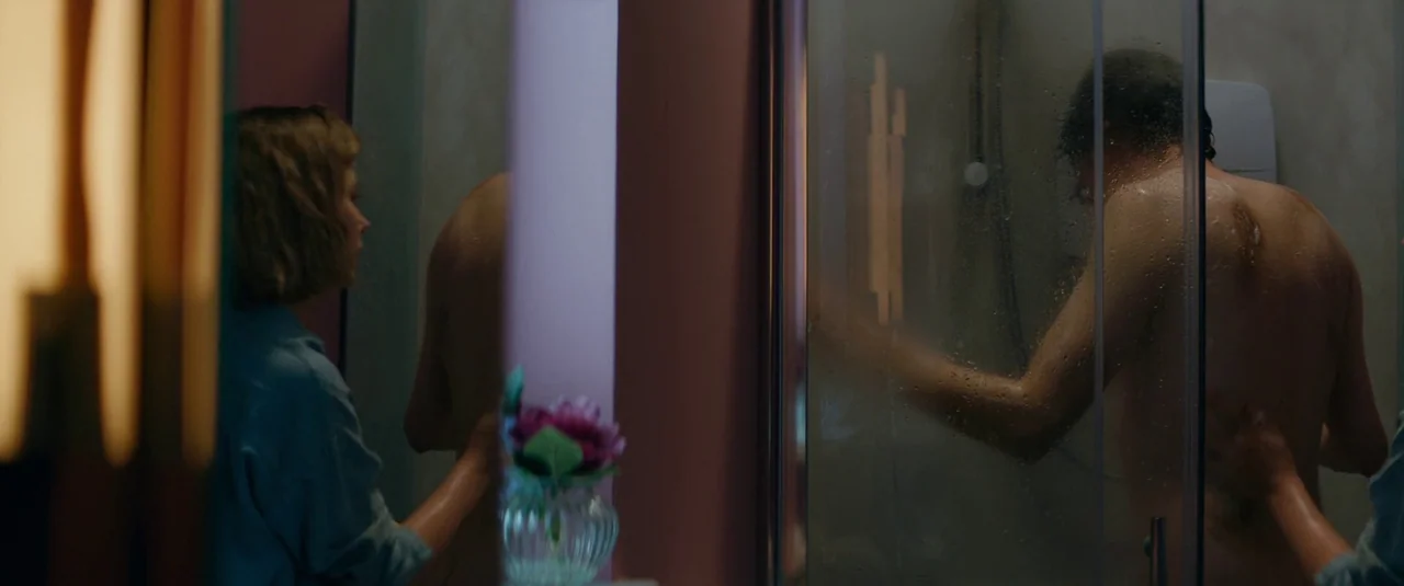 Vivarium (2019) movie still: medium — A dimly lit room features a woman standing in front of a mirror. She watches a person standing in a…