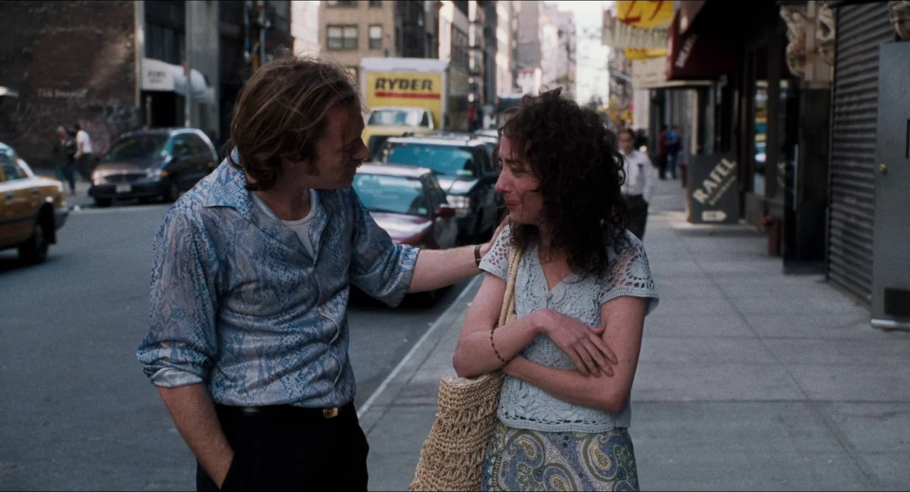Happiness (1998) movie still: medium — A city street sidewalk features parked cars and urban storefronts in the background. A man stands beside a…
