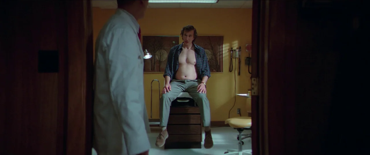 White Noise (2022) movie still: wide — A medical exam room features yellow walls and hanging wall art behind a patient. An adult male sits shirtless…