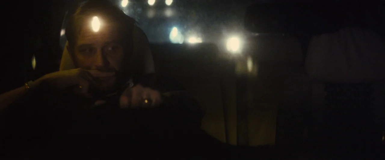 Locke (2013) movie still: medium — A man sits behind the steering wheel of a car at night. Outside lights reflect off his forehead while the cabin…