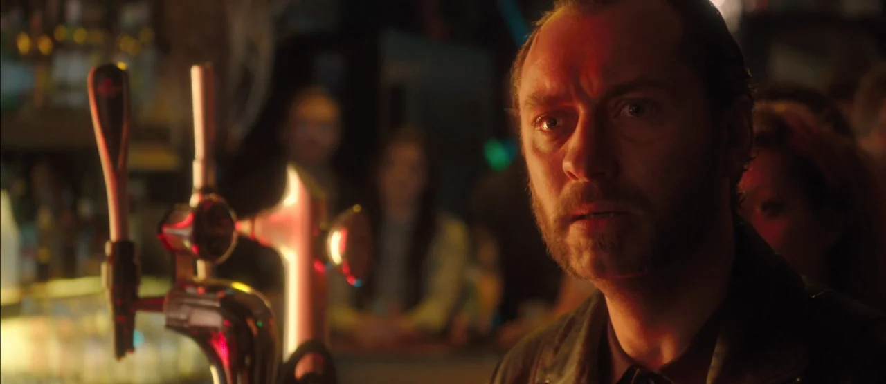 Dom Hemingway (2013) movie still: closeup — A dimly lit bar with metal beer taps in the foreground and blurry patrons. A man with a trimmed beard…
