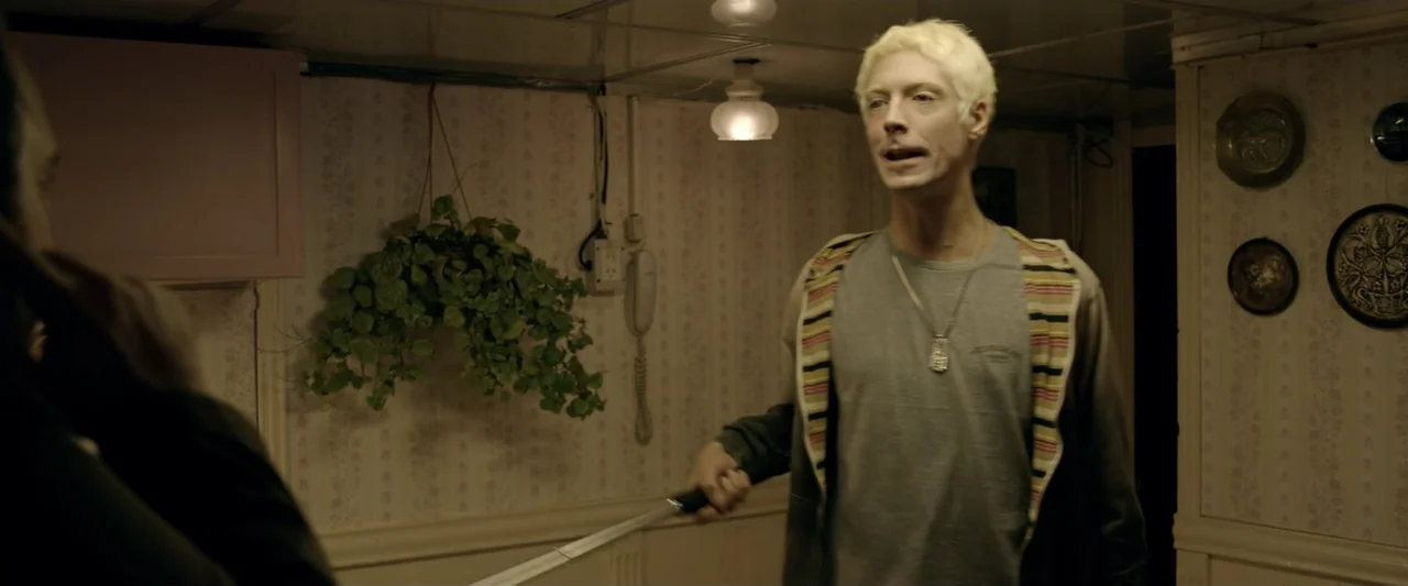 Perdida (2018) movie still: medium — A dimly lit room features floral wallpaper and a hanging plant on the wall. A blond man holds a sword while…