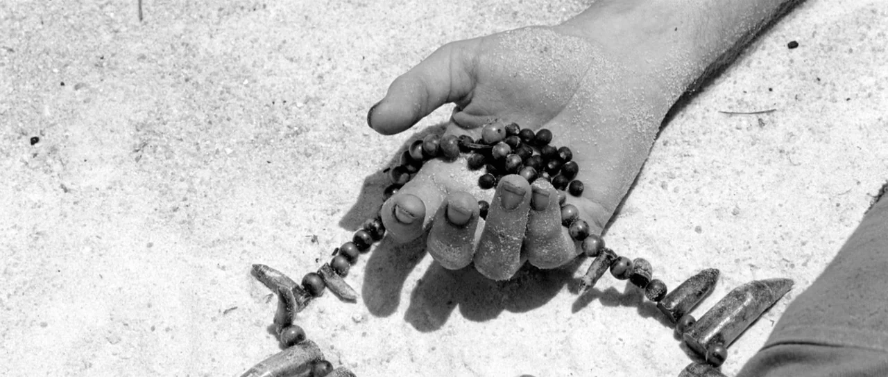Embrace Of The Serpent (2015) movie still: closeup — A human hand lies open on coarse light sand, coated in fine granules. The palm holds dark beads…