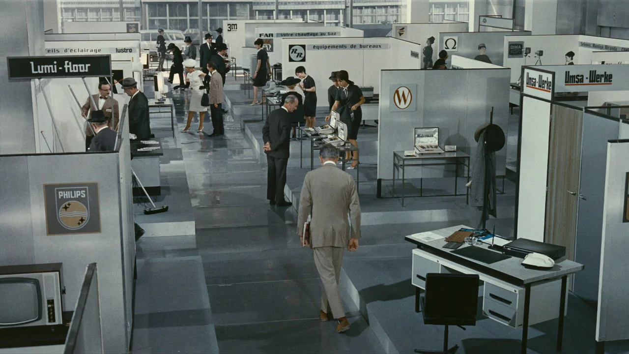 Playtime (1967) movie still: wide — An industrial trade show floor features partitioned booths displaying office equipment and consumer goods.…