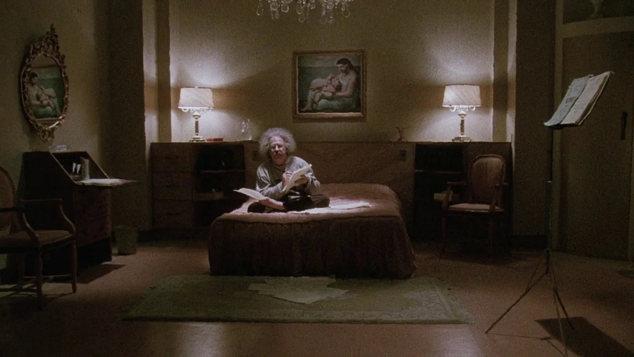 Insignificance (1985) movie still: wide — A dimly lit room features a central bed with a man sitting on it. Two bedside lamps frame the bed while a…
