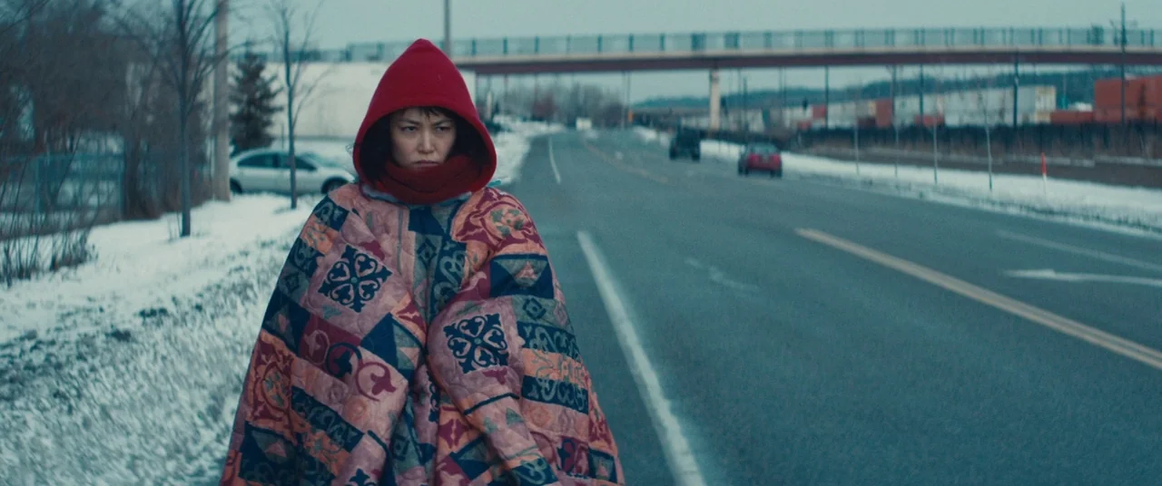 Kumiko the Treasure Hunter (2014) movie still: medium — A snowy roadside with a road stretching toward an overpass in the background. A woman walks…