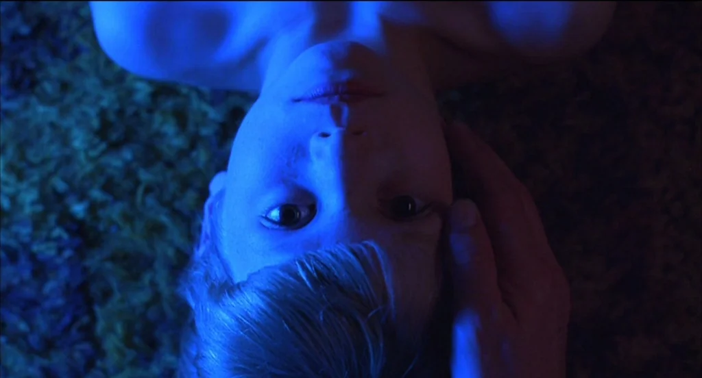 Mysterious Skin (2004) movie still: closeup — A child lies on a textured, dark-colored carpet indoors at night. An adult hand reaches in to touch…