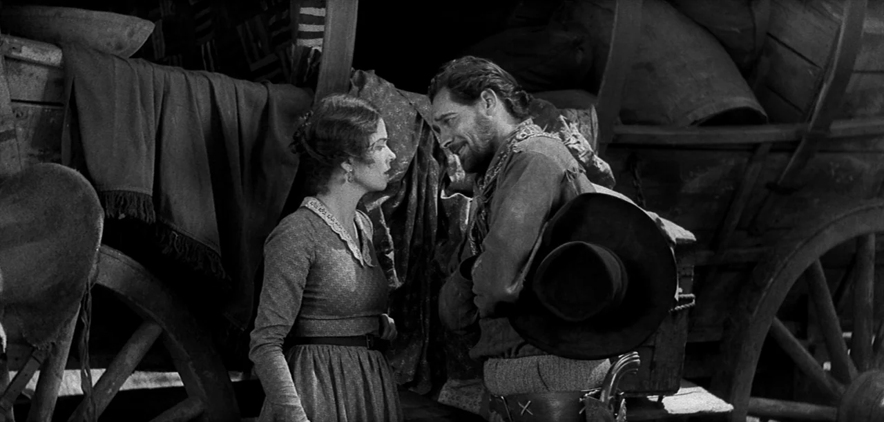 The Big Trail (1930) movie still: medium — A man and woman stand near a large wooden covered wagon with fabric draped over. They engage in a…
