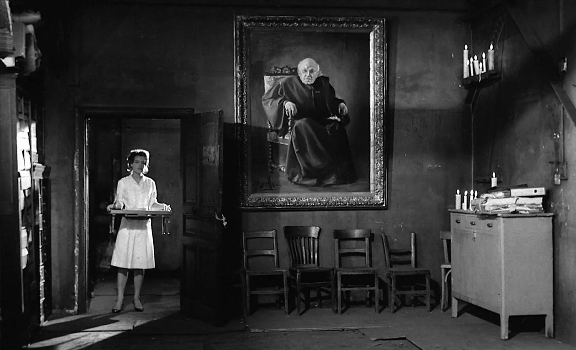 The Trial (1962) movie still: fullbody — A dimly lit room contains wooden chairs and a large portrait hanging on the wall. A woman in a white dress…