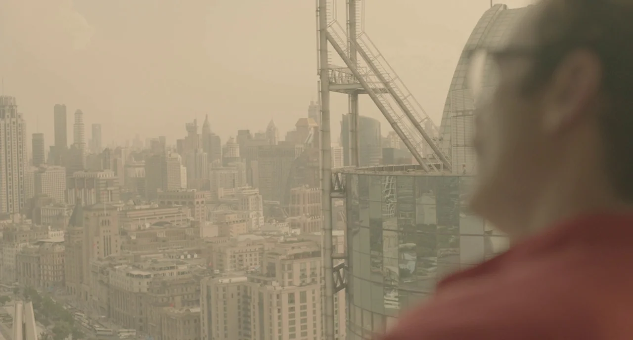 Her (2013) movie still: medium — A high-angle view looks over a densely packed urban skyline from a rooftop. A man in a red shirt stands in the…