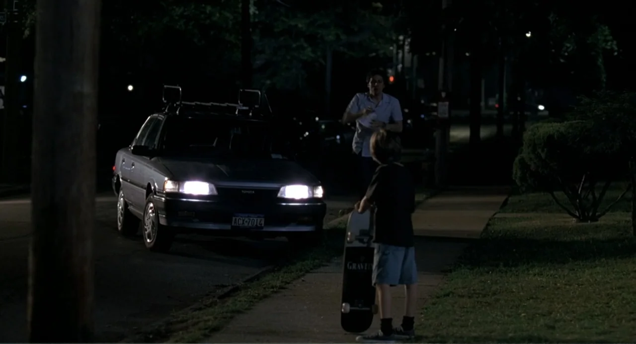 Mi Perfecto Ex (2024) movie still: medium — A suburban sidewalk at night is illuminated by the headlights of a parked car. A man approaches a child…