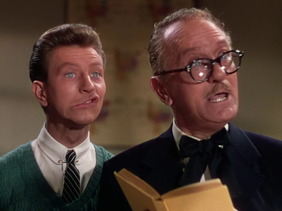 Singin’ in the Rain (1952) movie still: medium — A brightly lit indoor classroom space with a map on the wall. An older man wearing a bowtie holds a…