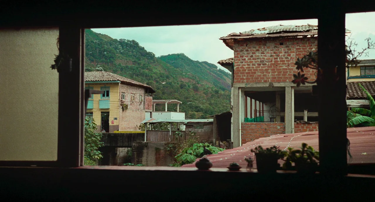 Memoria (2021) movie still: establishing — View through a dark window frame looking out over small hillside houses made of brick. A lush green…